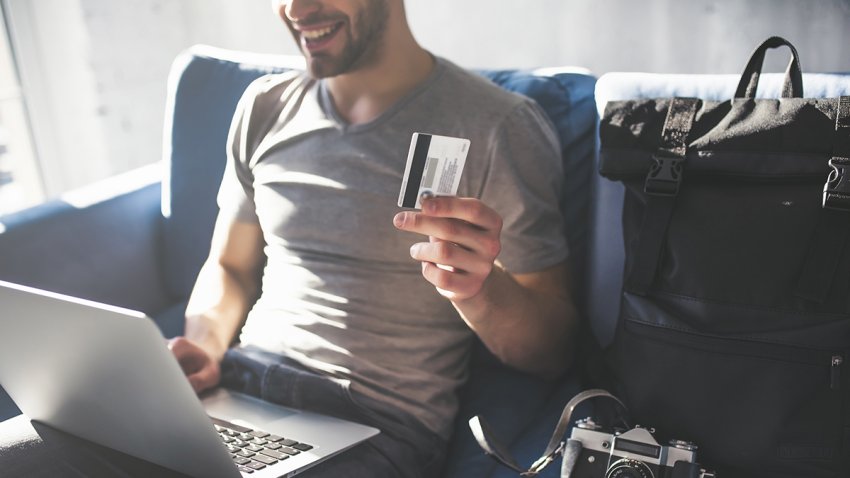 Handsome young man is sitting at home with laptop and credit card in hands. Preparing for traveling